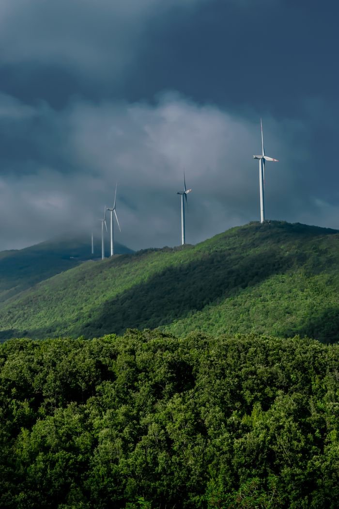 services-03 Series of wind turbines on a green mountain range under a dramatic cloudy sky.